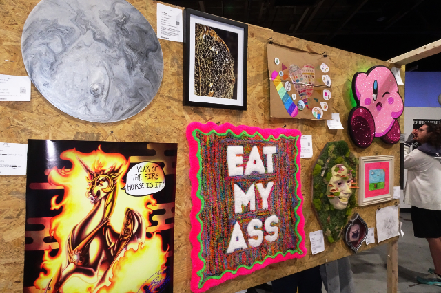 A temporary wall covered in artwork in a range of scale and media. Most notably: A brigh, neon punch rug stating EAT MY ASS, an enamel circle with marbled ink on the surface, resembling the moon, a giant cut-out of the video game character Kirby, covered in glitter, and an inkjet print on large-format paper of Princess Celestia from My Little Pony, surrounded by dark flames.