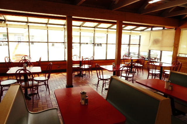 The sunlit interior of a fast food restaurant, with rows of brightly colored booths, chairs arranged around small tables, and large terrarium windows.