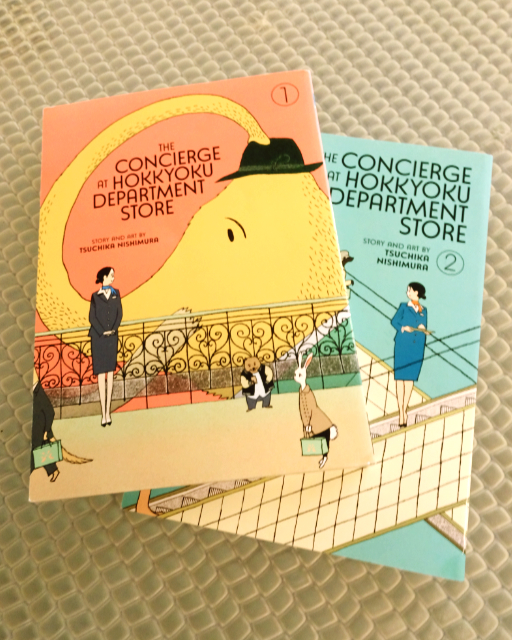 Two volumes of the manga series The Concierge at the Hokkyoku Department Store sitting on a textured glass table.