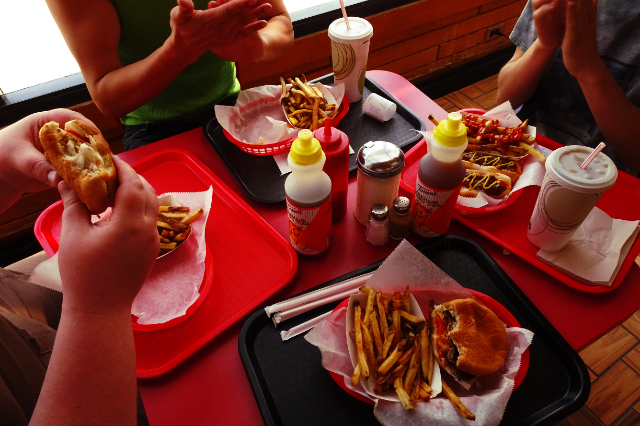 looking down at a table full of freshly-prepared burgers and hot dogs, with hands reaching for sandwiches, soda cups and iced tea bottles.