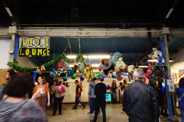 A large warehouse full of people, with an art installation of many foil balloons hanging from the ceiling. There is a large handmade sign that reads VIDEO LOUNGE over everyone's heads.